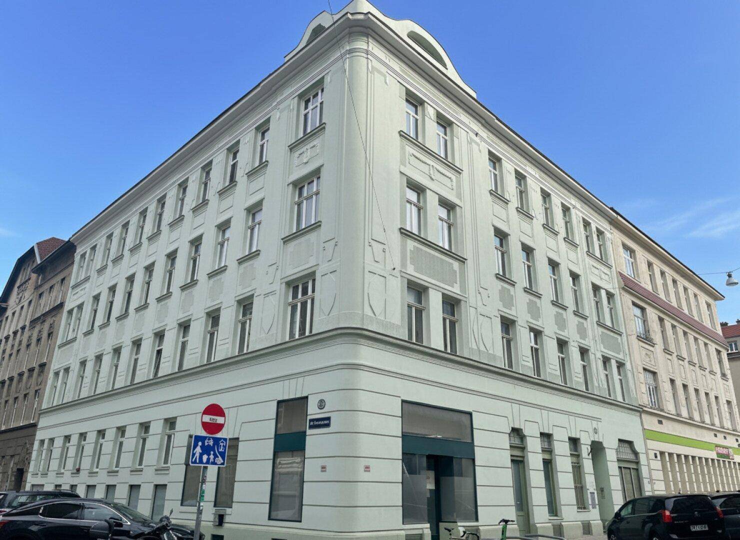 Historisches Eckgebäude mit vielen Fenstern und einer hellgrünen Fassade unter blauem Himmel.