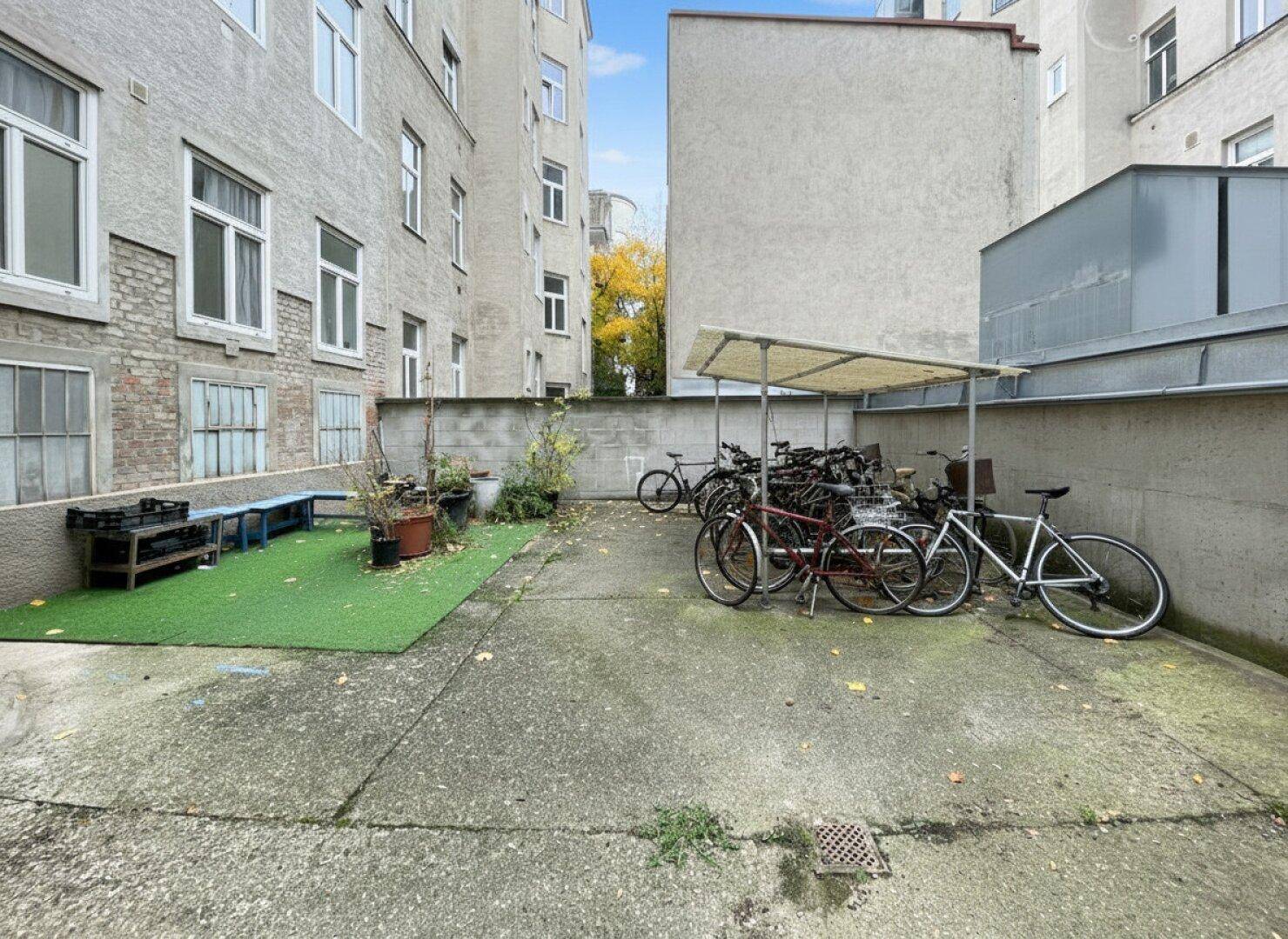 Hinterhofbereich mit Fahrradabstellplatz, Kunstrasen und einer Mauer, umgeben von Gebäuden.