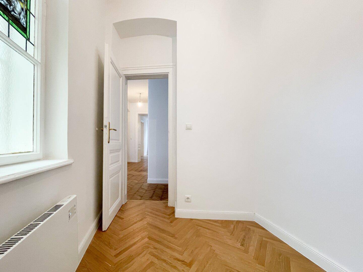Kleines Zimmer mit Fischgrätparkett, einem Buntglasfenster und einer offenen Tür, die in den Flur führt.