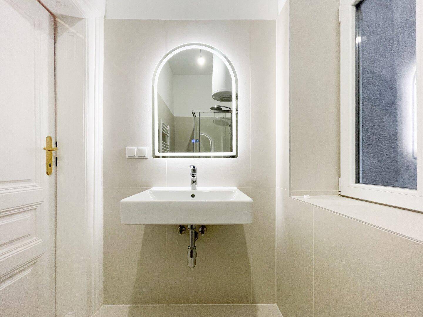 Modernes Badezimmer mit einem einzelnen Waschbecken, beleuchtetem Spiegel und hellen Fliesen.