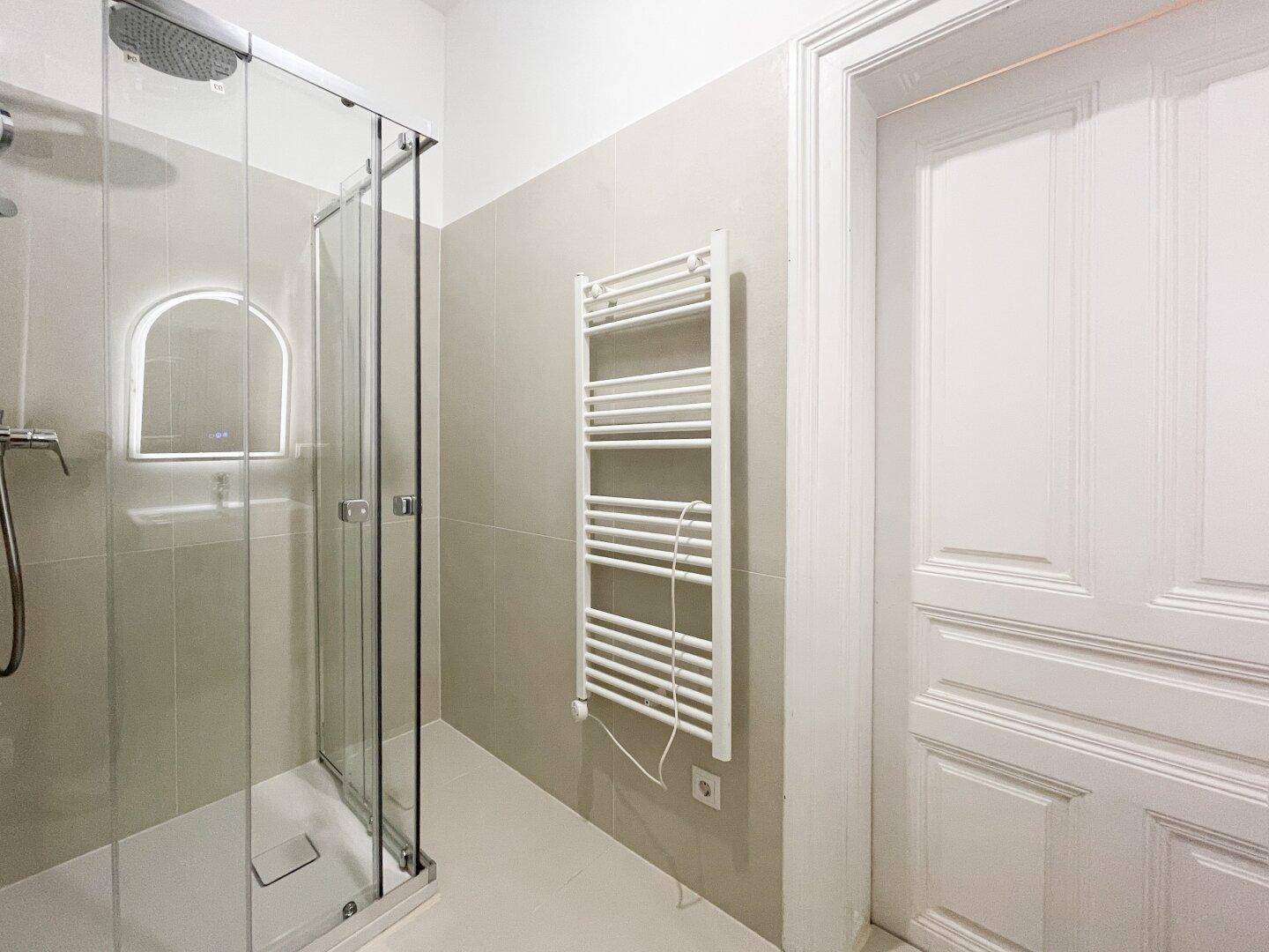 Modernes Badezimmer mit Glasduschkabine, beheiztem Handtuchhalter und hellen Wand- und Bodenfliesen.