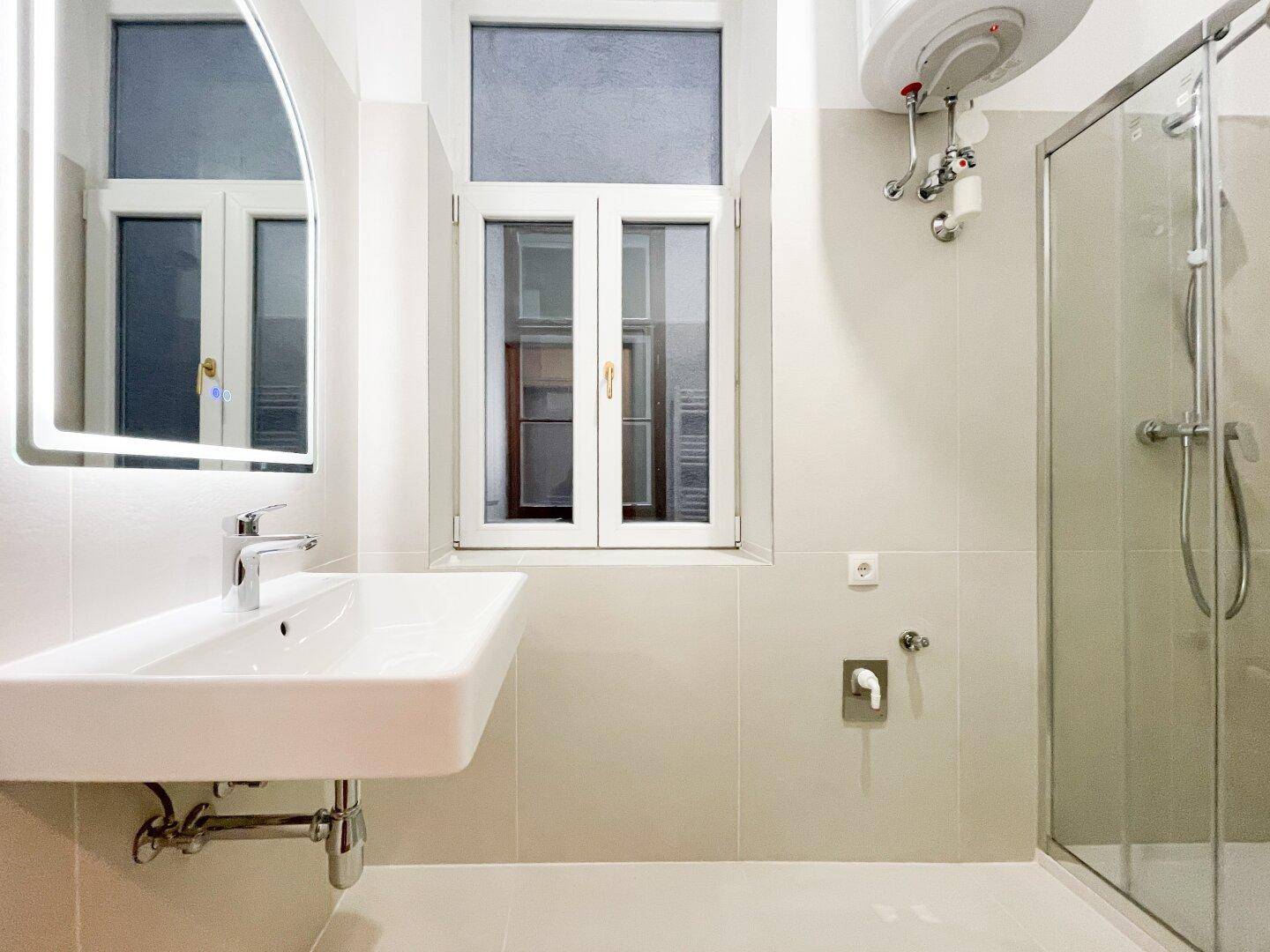 Modernes Badezimmer mit weißem Waschbecken, beleuchtetem Spiegel und einer Glasduschkabine.