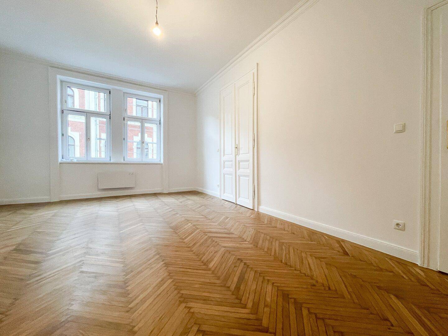 Wohnraum mit Fischgrätparkett, hohen Decken, großen Fenstern und einem praktischen Einbauschrank.