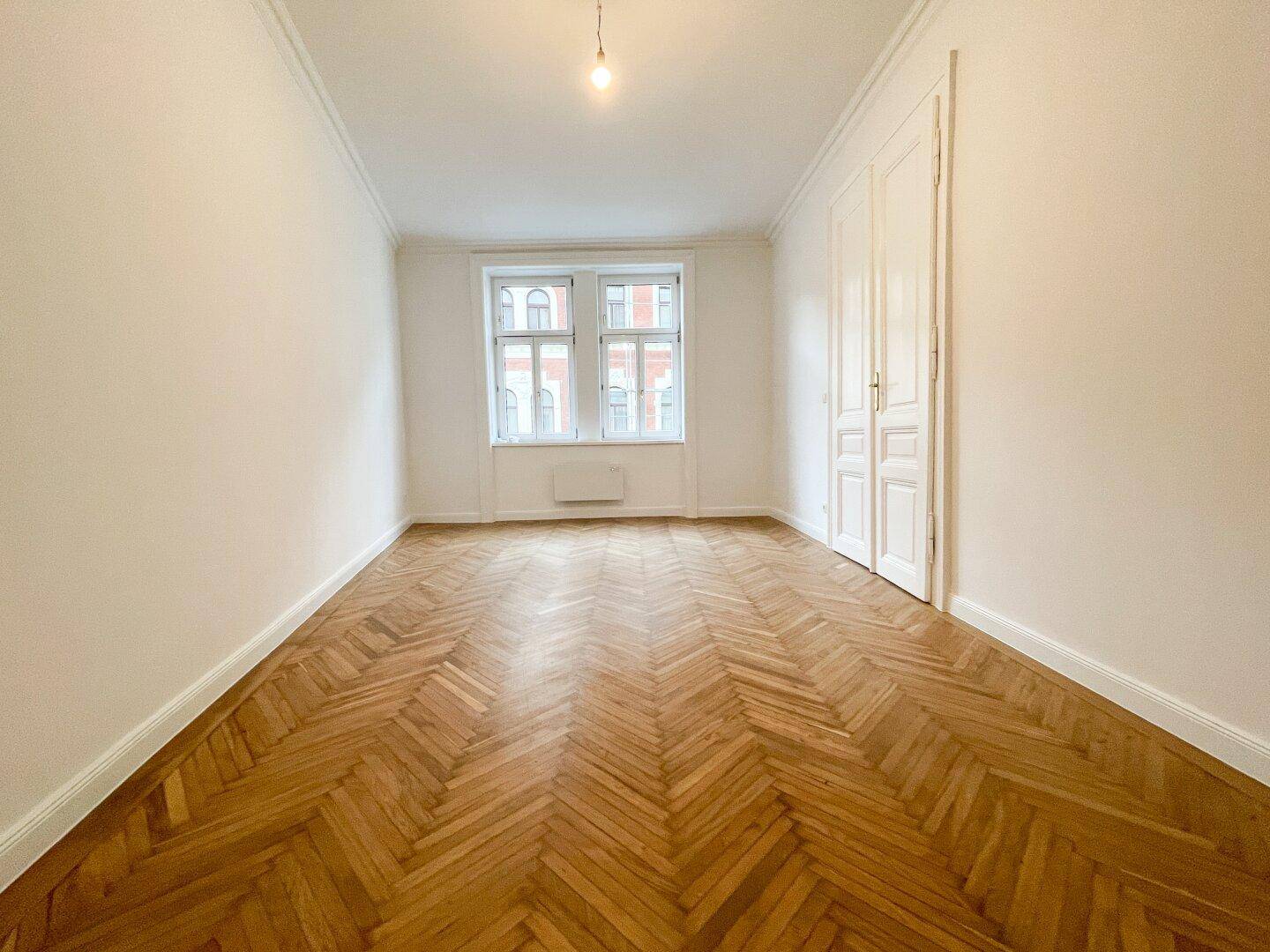 Langer Wohnraum mit klassischem Fischgrätparkett, hohen Decken und zwei Fenstern, die für viel Licht sorgen.