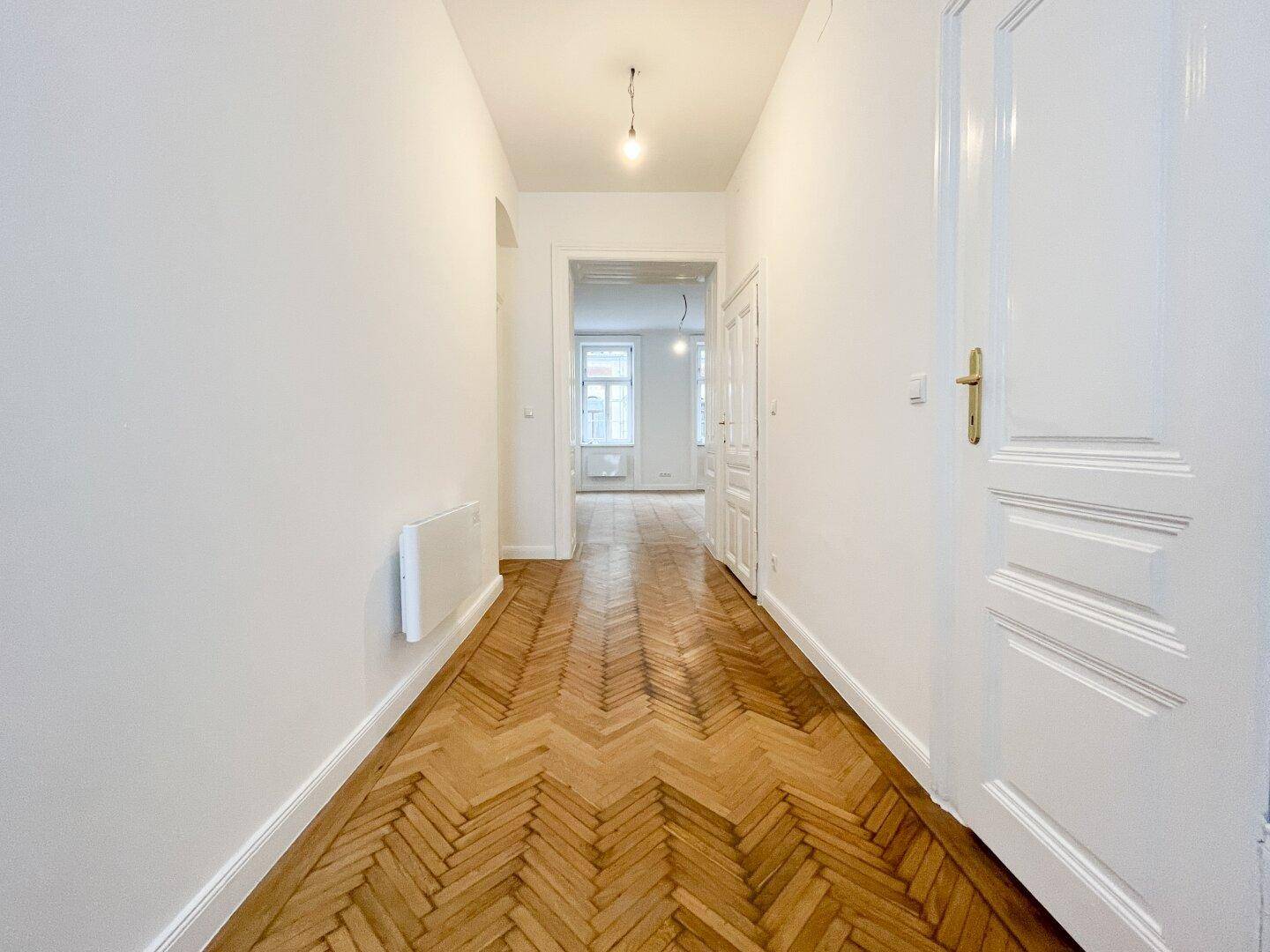Langer Flur mit elegantem Fischgrätparkett, weißen Wänden und hohen Decken, der zu weiteren Räumen führt.