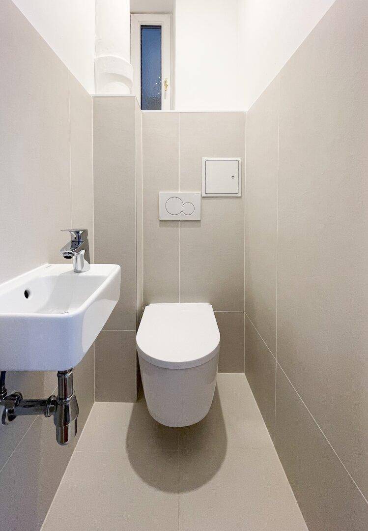 Kompaktes Gäste-WC mit wandhängendem WC, kleinem Waschbecken und hellen Fliesen.