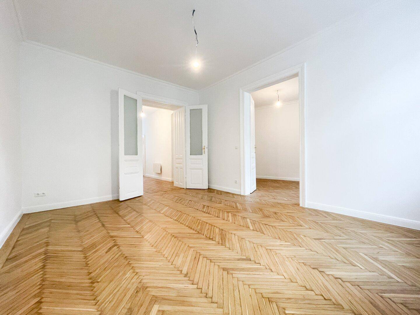 Helles Wohnzimmer mit Fischgrätparkett und hohen Decken, durch Doppeltüren mit weiteren Räumen verbunden.