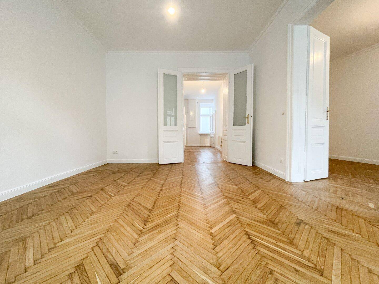 Geräumiges Wohnzimmer mit elegantem Fischgrätparkett und hohen Decken, das durch offene Türen weitere Räume zeigt.