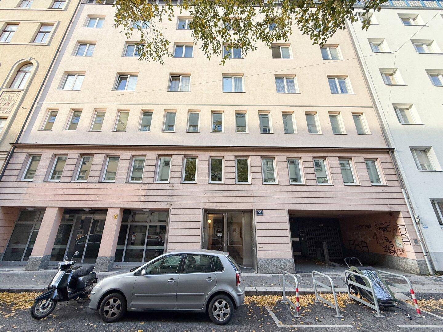 Die Fassade des Mehrfamilienhauses mit mehreren Etagen und Fenstern, davor parkende Fahrzeuge.