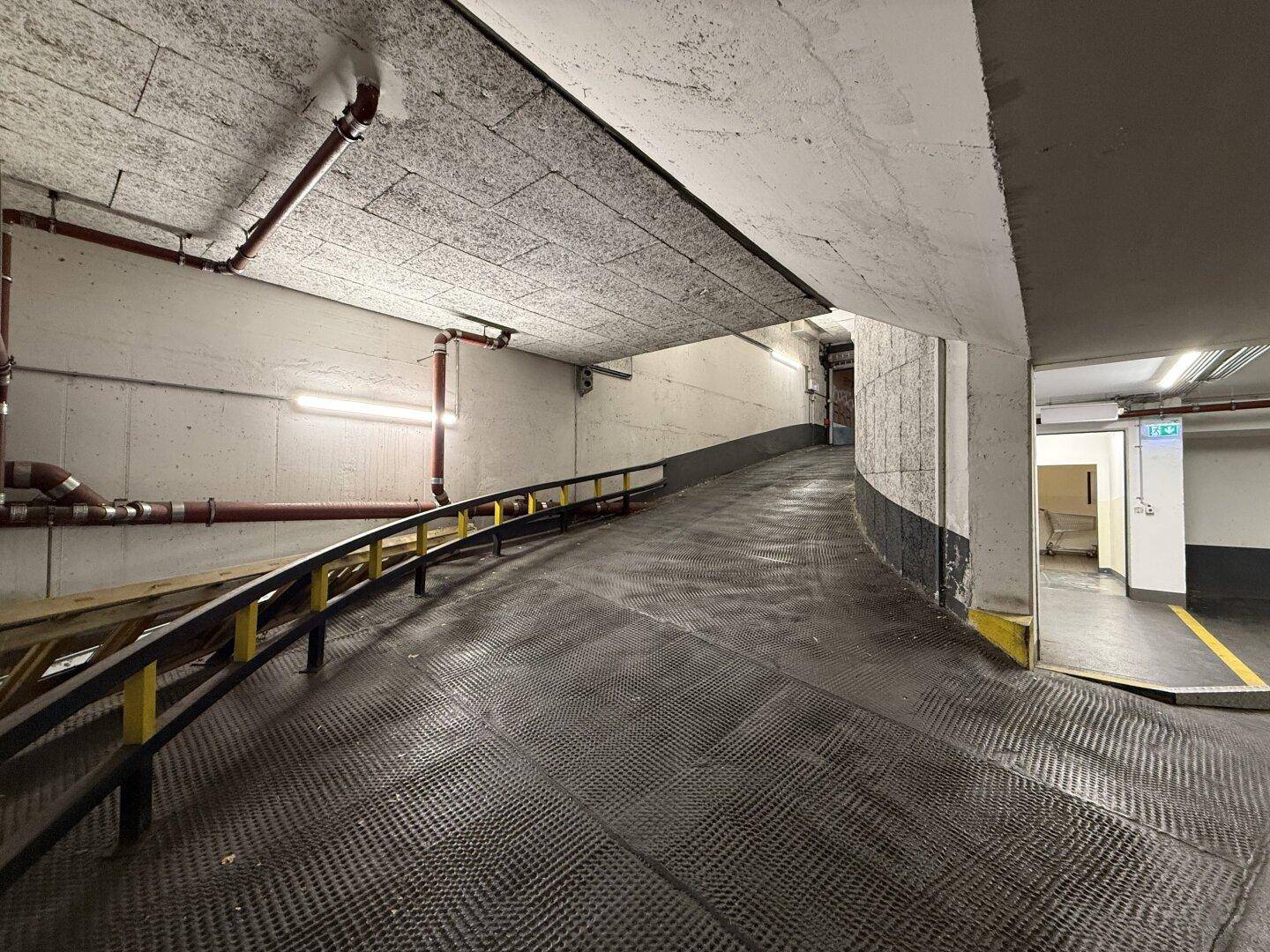 Die Tiefgaragenrampe mit Handlauf und sichtbaren Rohrleitungen an der Decke und Wand.