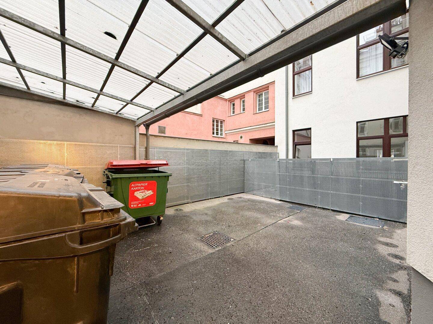 Überdachter Außenbereich mit Mülltonnen und Blick auf die umliegenden Gebäude.