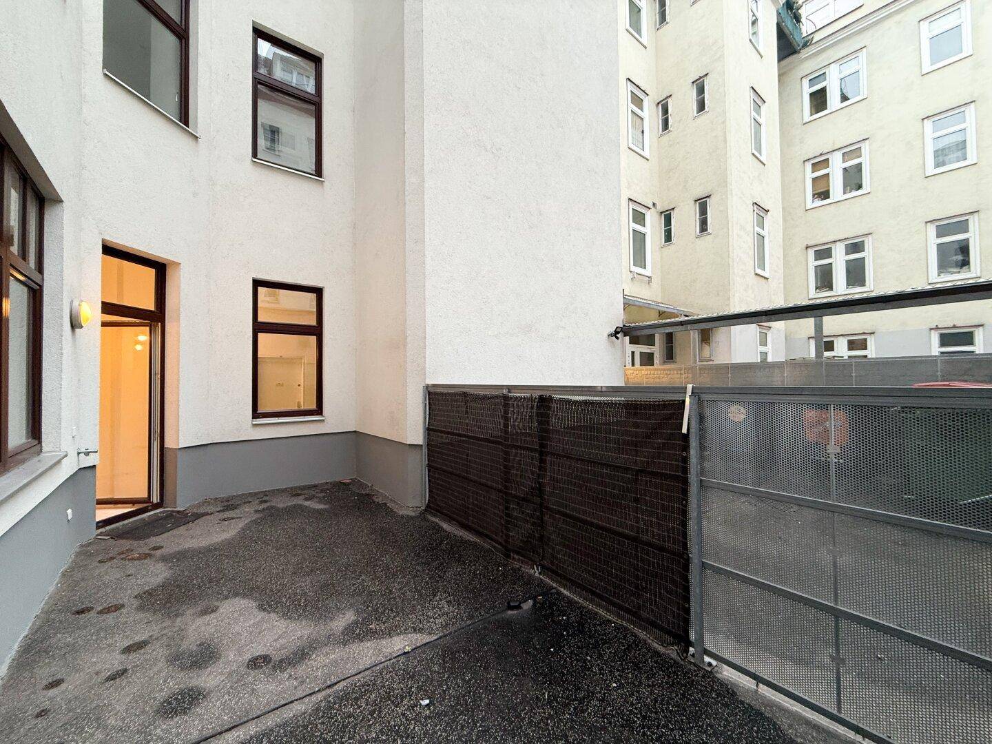Innenhof mit gepflastertem Boden und einer Metallabtrennung, umgeben von Gebäuden.