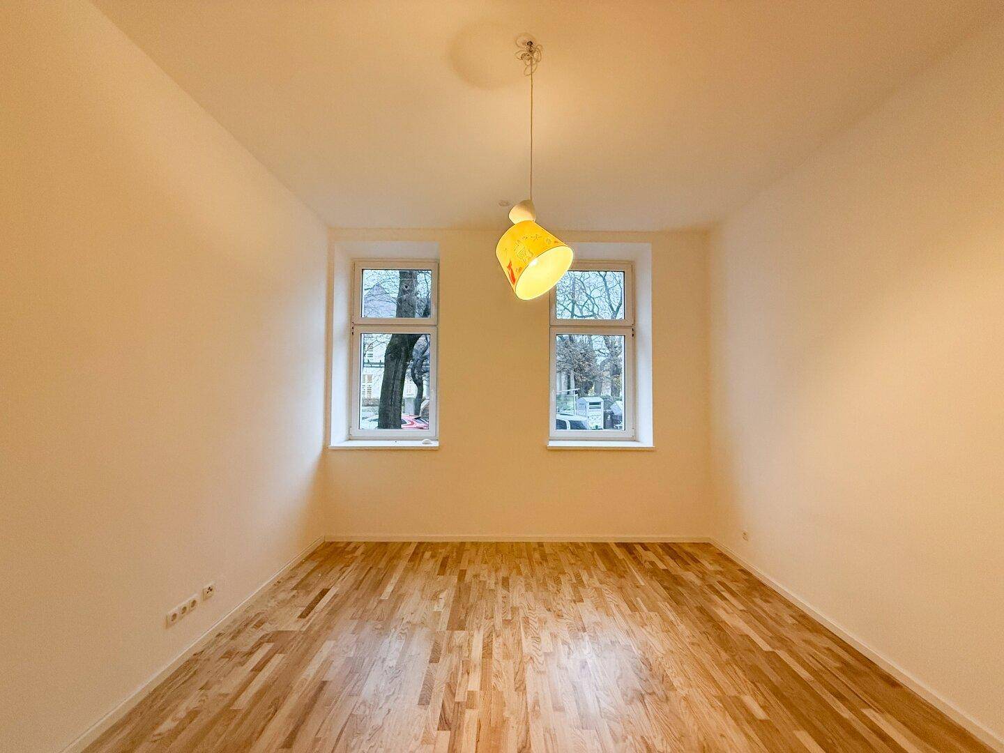 Leeres Wohnzimmer mit hellem Parkettboden und viel Tageslicht durch zwei Fenster.