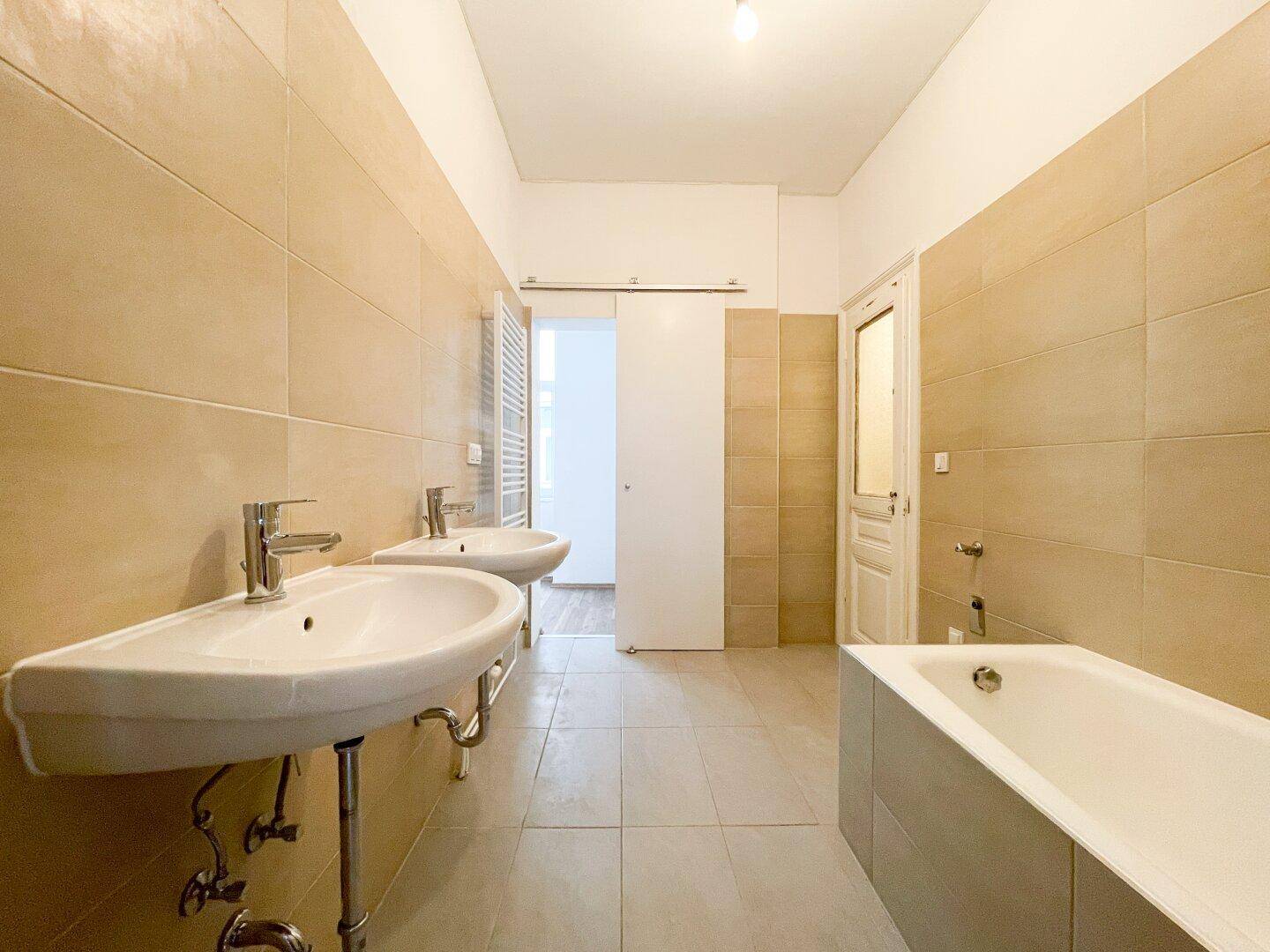 Geräumiges Badezimmer mit Doppelwaschbecken, Badewanne und beigefarbenen Fliesen.