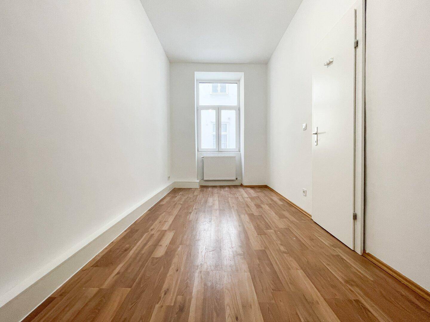 Schmaler, heller Raum mit Holzboden und einem Fenster, ideal als Schlaf- oder Arbeitszimmer.