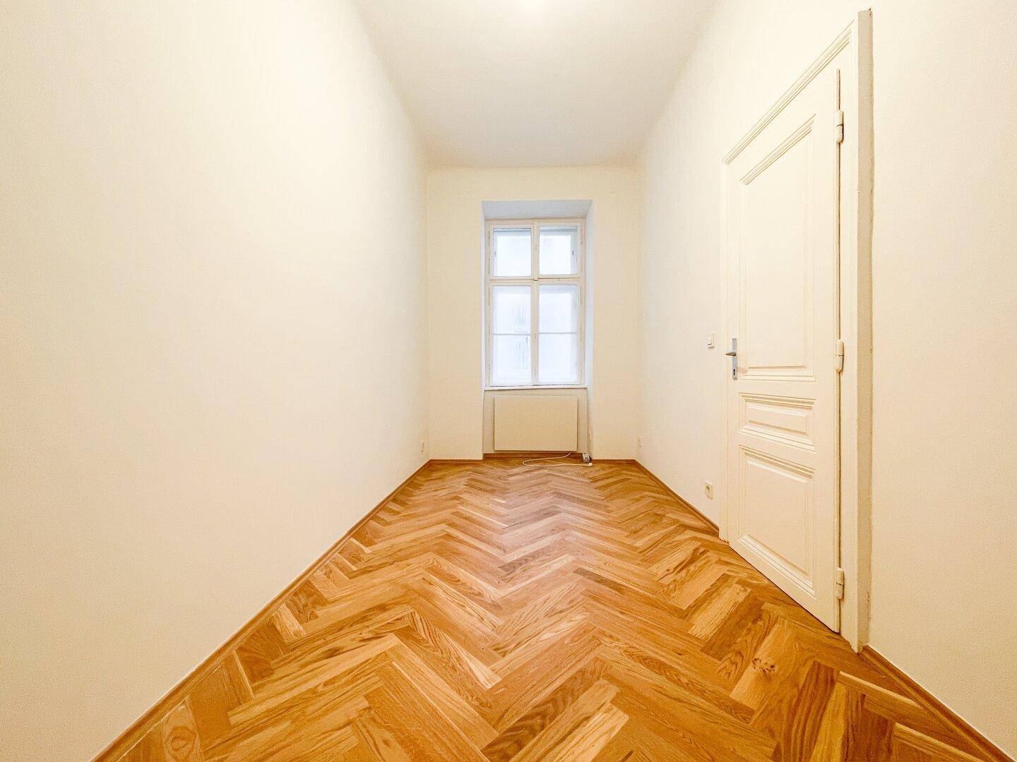 Langer, heller Raum mit Fischgrätparkett und einem Fenster, geeignet als Schlaf- oder Arbeitszimmer.