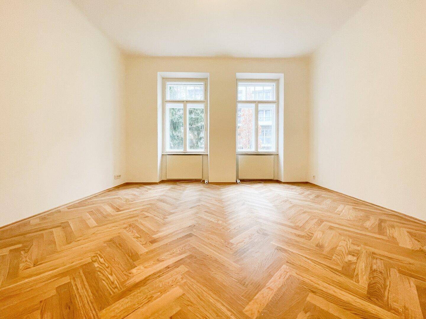 Heller, leerer Raum mit Fischgrätparkett und zwei großen Fenstern, ideal als Wohnzimmer.