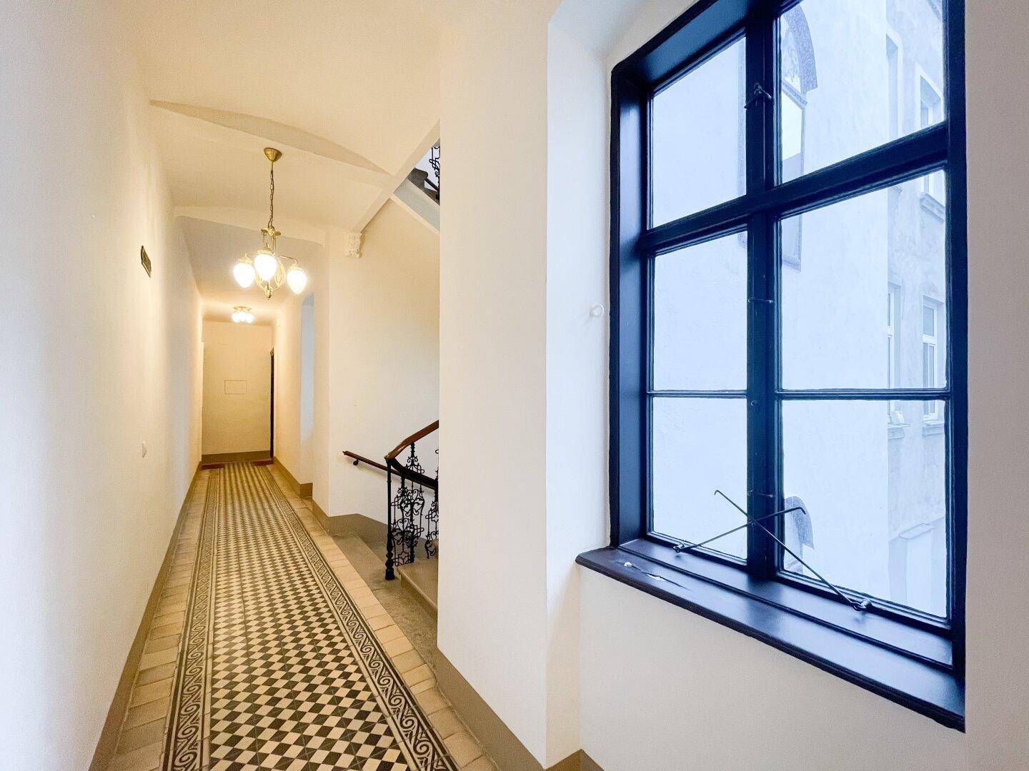 Helles Treppenhaus mit großem Fenster, Fliesenboden und Blick in den langen Gang des Altbaus.