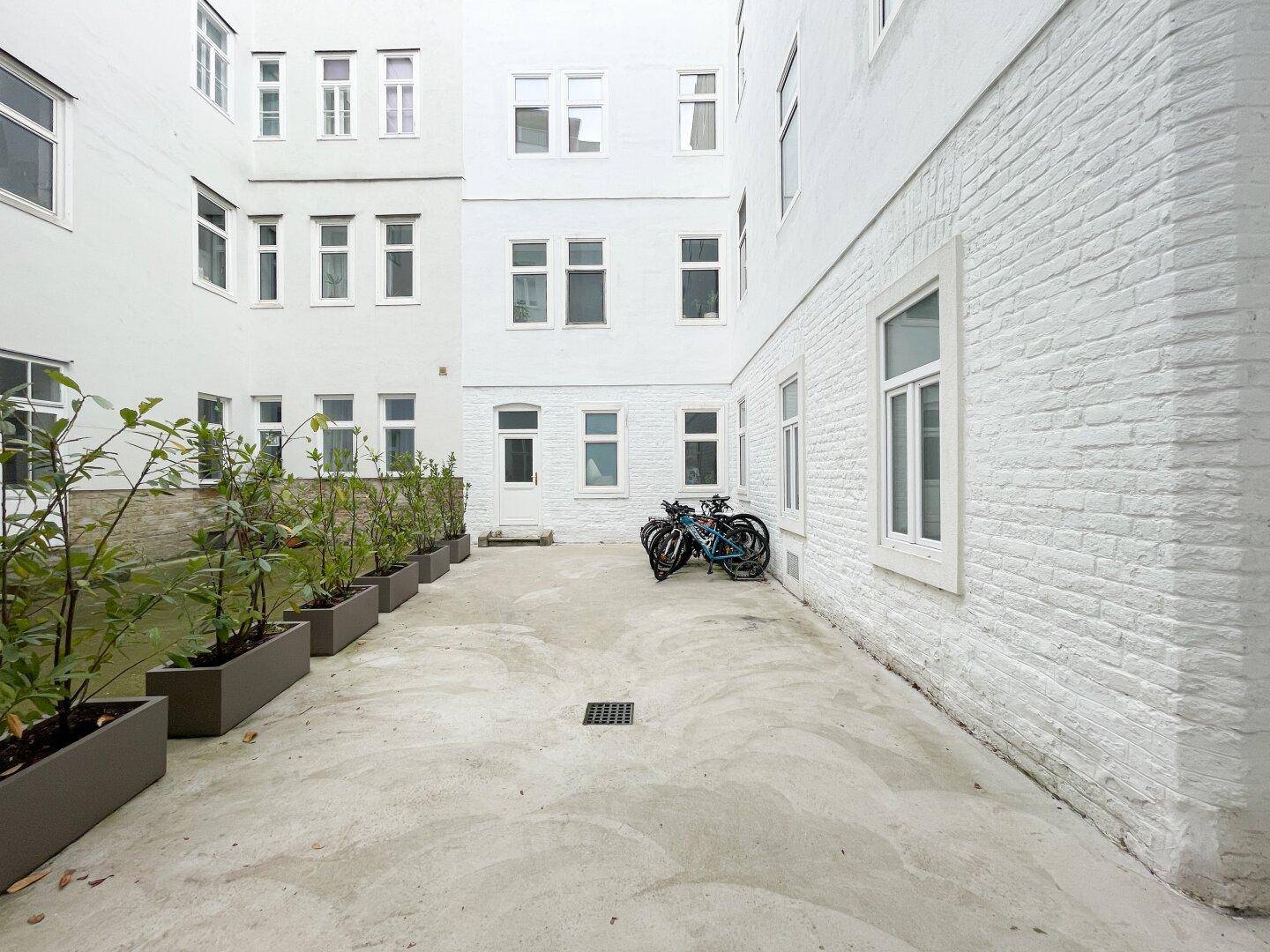 Weiß gestrichener Innenhof eines Gebäudekomplexes mit Pflanzenkübeln und Fahrradabstellplatz.
