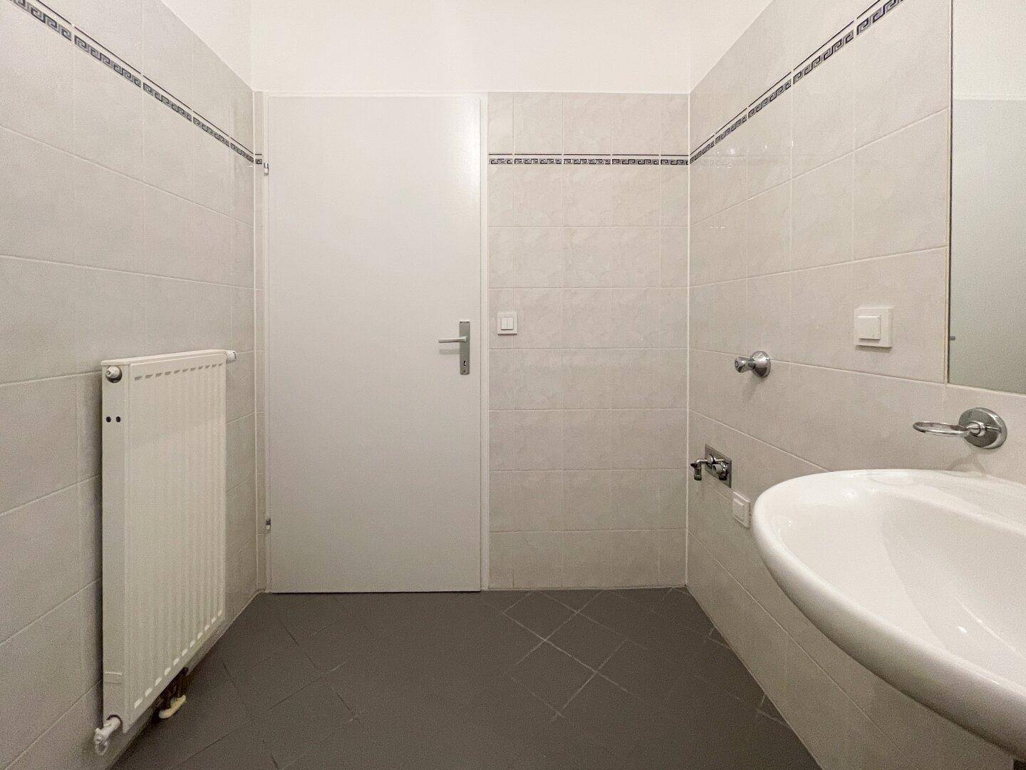 Badezimmer mit Waschbecken, Heizkörper und hellen Fliesen, bietet grundlegende Ausstattung.