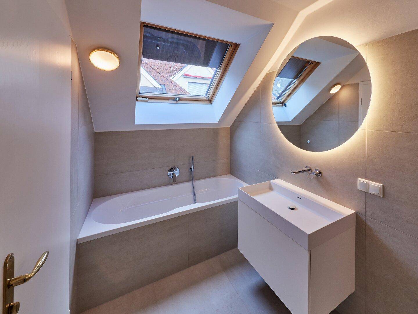 Modernes Badezimmer mit Badewanne, Waschtisch, beleuchtetem Spiegel und Dachfenster unter einer Schräge.