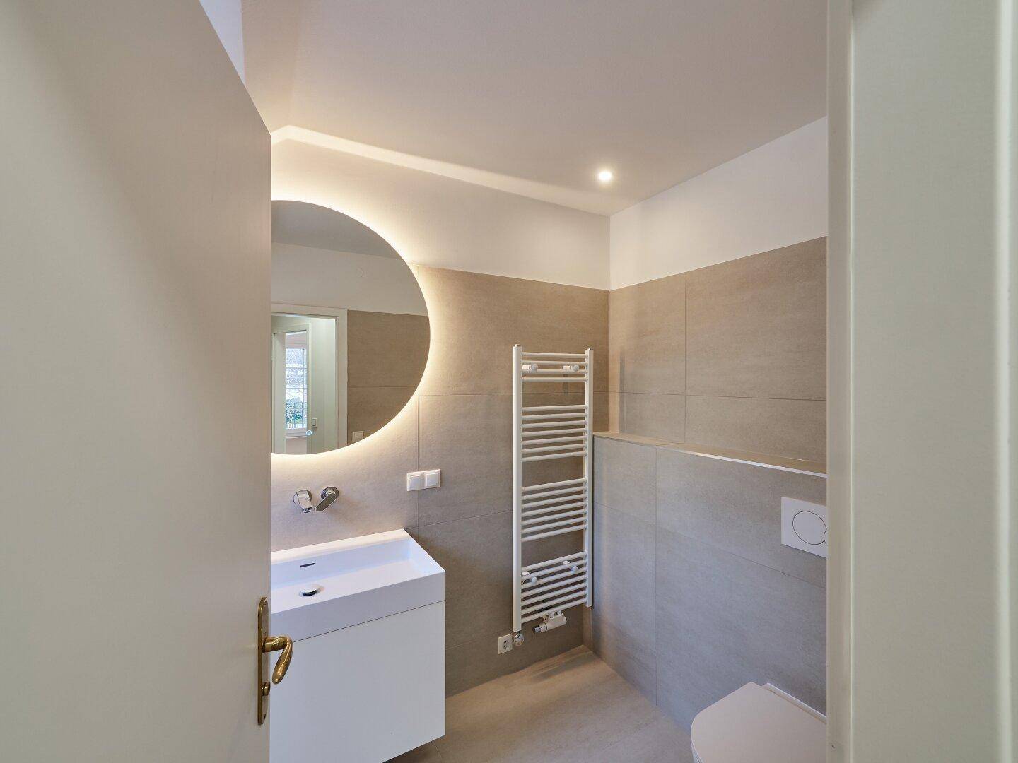 Elegantes Badezimmer mit modernem Waschtisch, WC, beleuchtetem Spiegel und beheiztem Handtuchhalter.