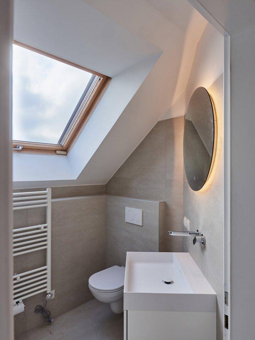 Modernes Gäste-WC mit Waschtisch, WC, beleuchtetem Spiegel und Dachfenster unter einer Schräge.