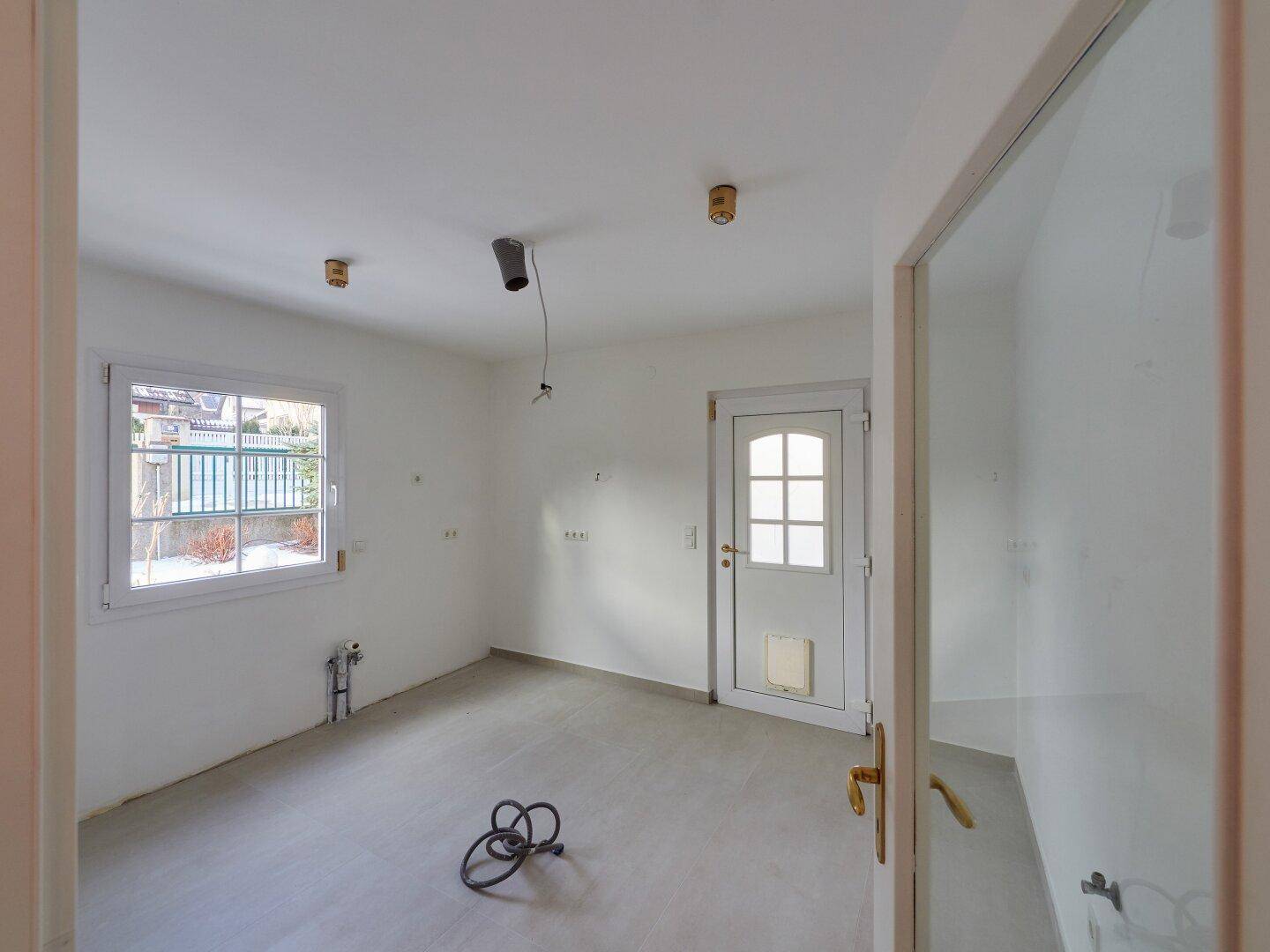 Leerer Raum mit Fliesenboden, Fenster und Anschlüssen, bereit für die Installation einer Küche oder als Hauswirtschaftsraum.