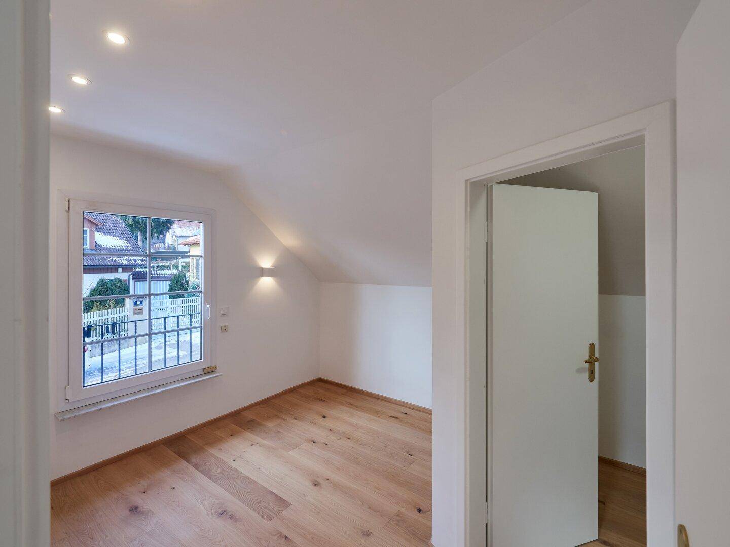 Helles Schlafzimmer mit Holzboden, Dachschräge und einem Fenster, das viel Tageslicht hereinlässt.