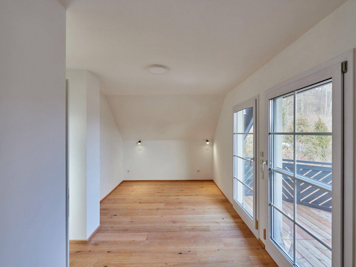 Helles Zimmer mit Holzboden, Dachschrägen und großen Fenstern mit Zugang zu einem Balkon.