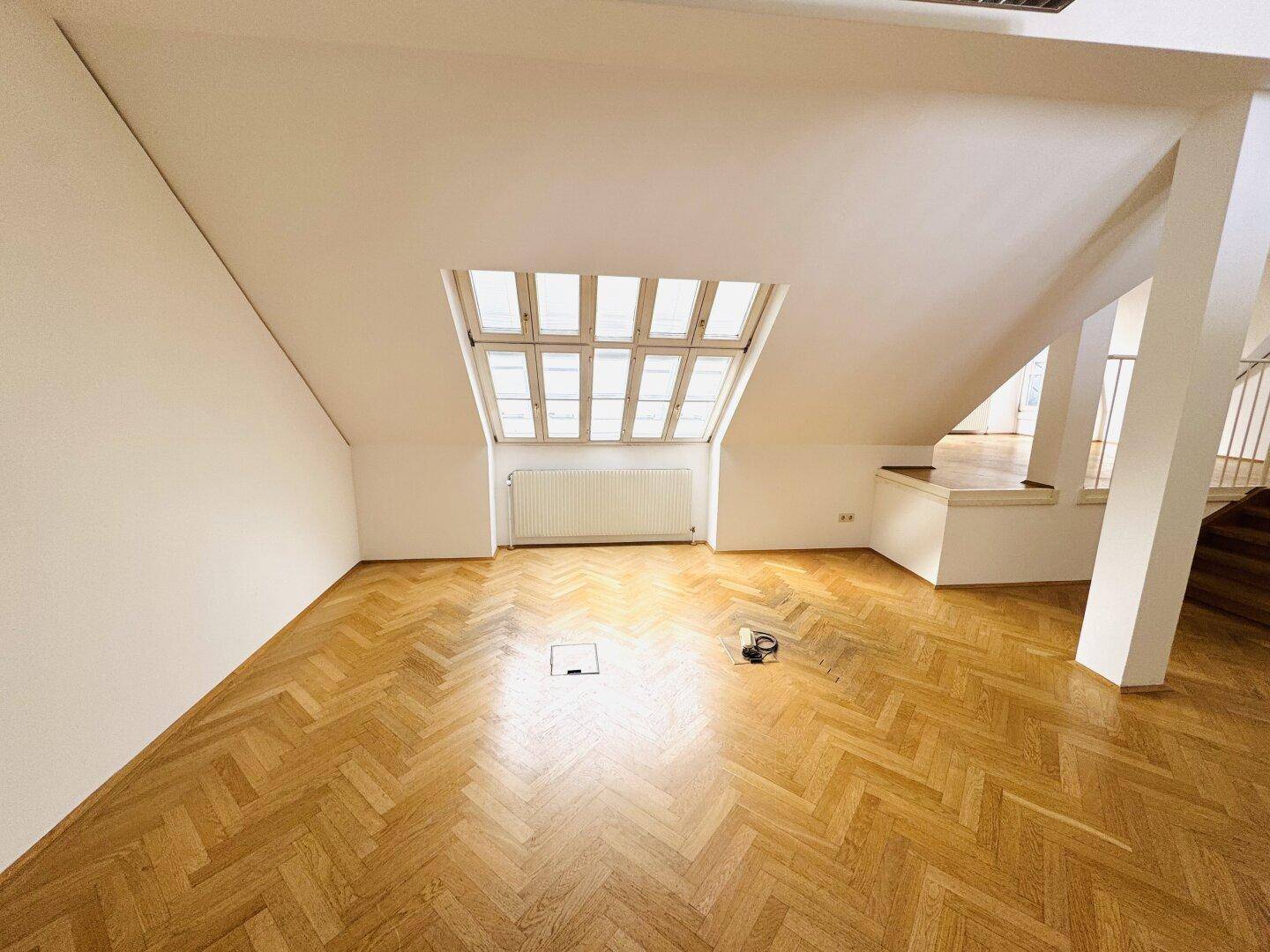 Geräumiger Raum im Dachgeschoss mit Holzparkett, Dachfenster und Heizkörper an der Wand.