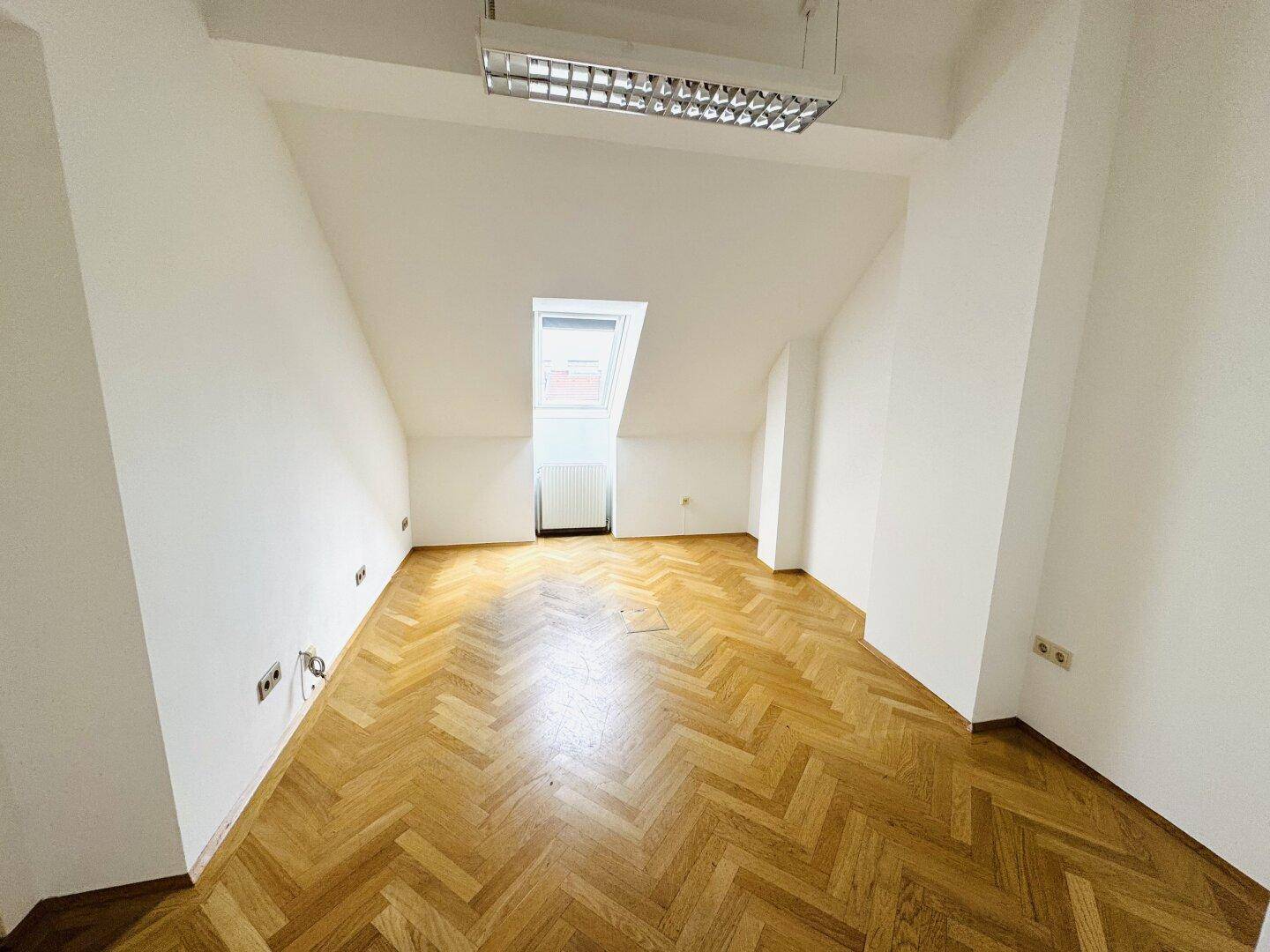 Dachgeschossraum mit Fischgrätparkett, Dachfenster und schrägen Wänden.