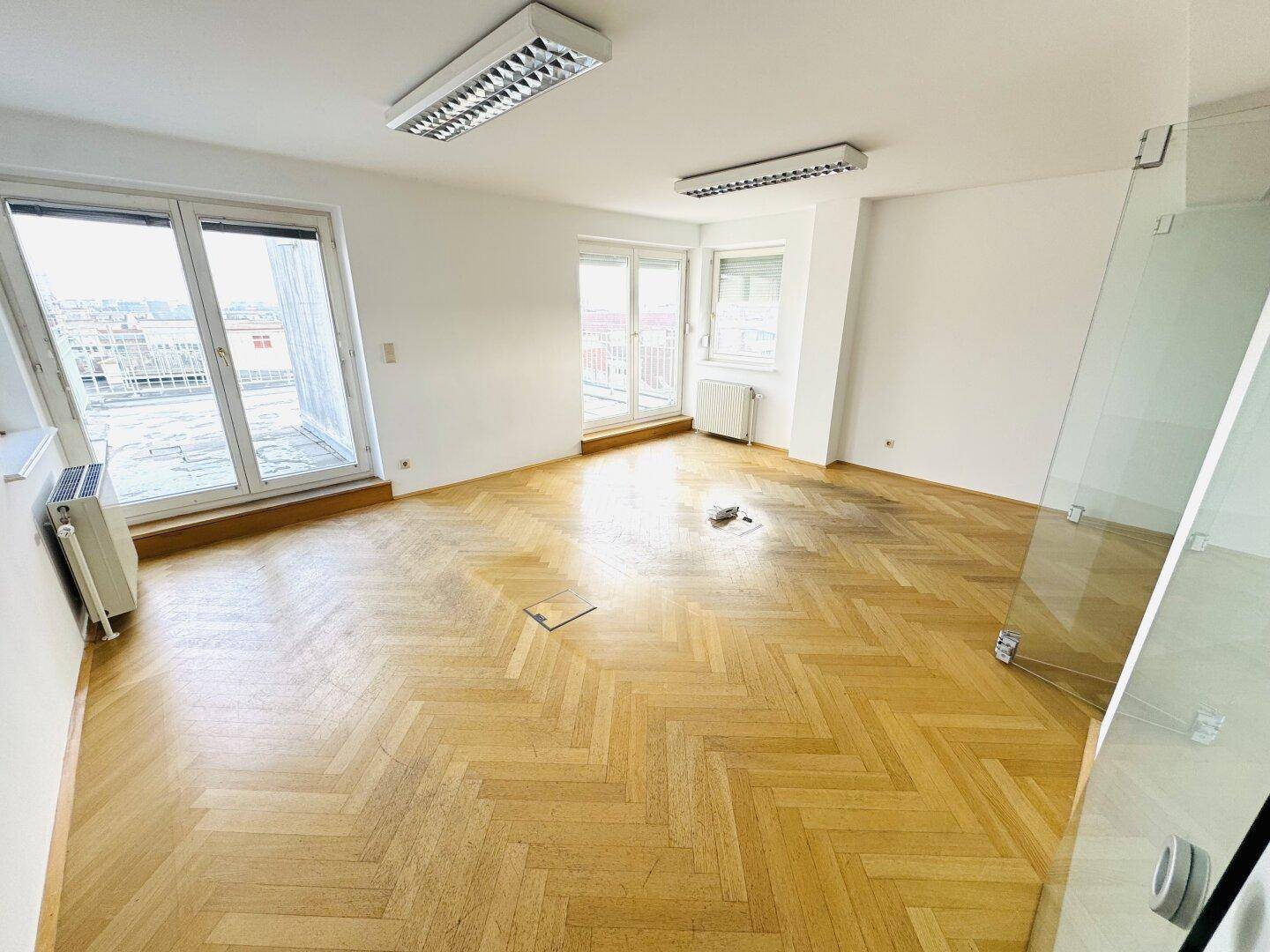 Großer Raum mit Fischgrätparkett, mehreren Fenstern und Blick auf die umliegenden Dächer.