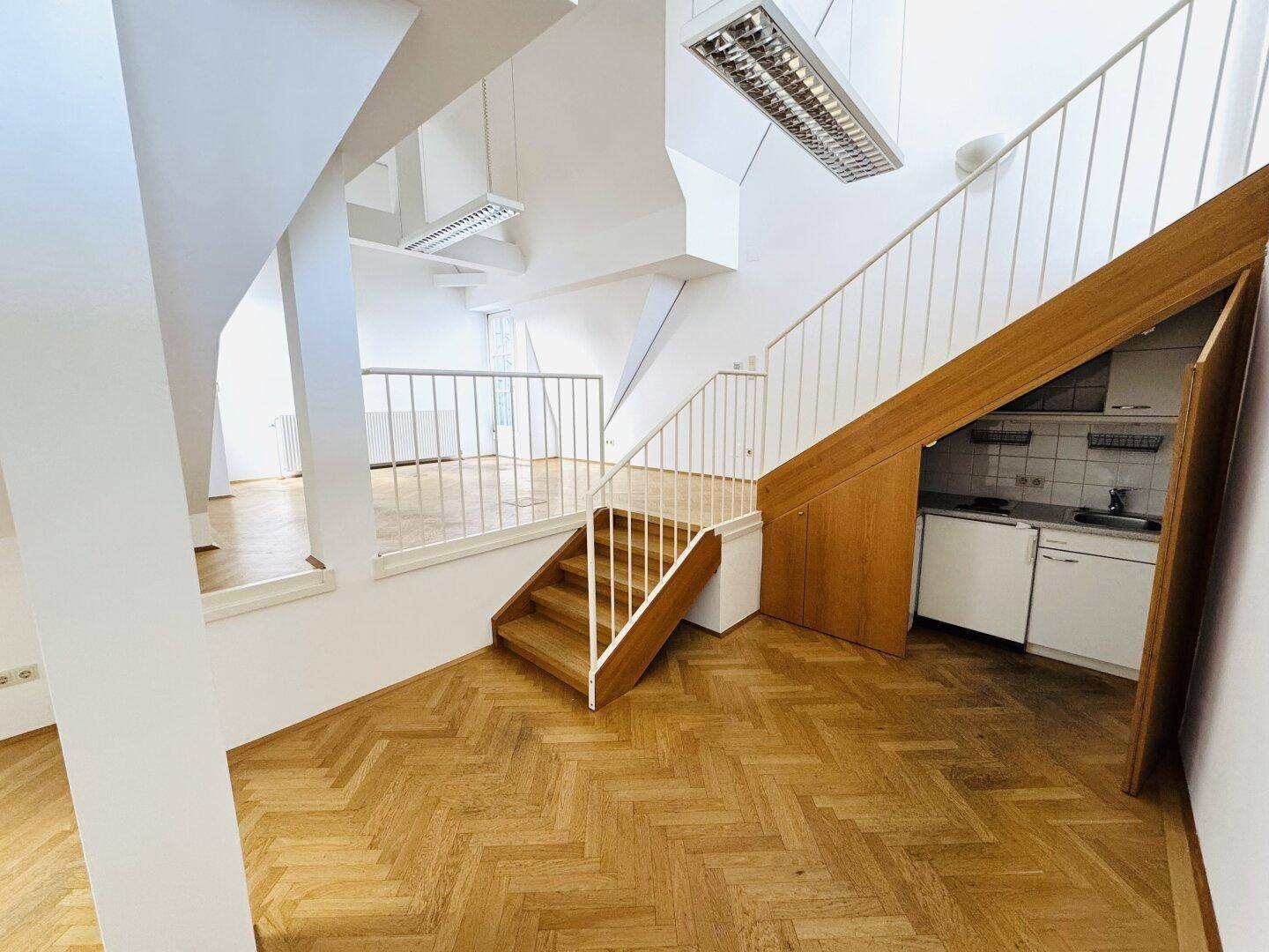 Heller Wohnbereich mit Holzparkett, offener Treppe und integrierter Küchenzeile unter der Treppe.