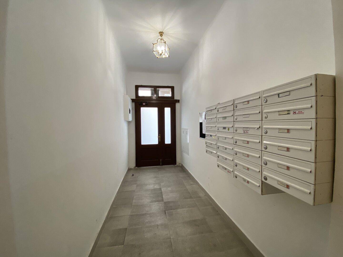 Heller Eingangsbereich eines Mehrfamilienhauses mit Briefkästen und einer Holztür am Ende des Flurs.