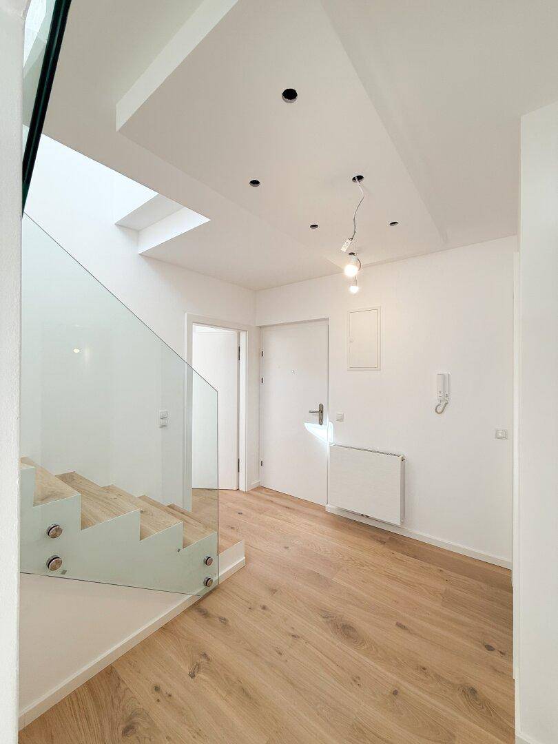 Moderner Eingangsbereich mit heller Holztreppe, Glasgeländer und Einbaustrahlern an der Decke.