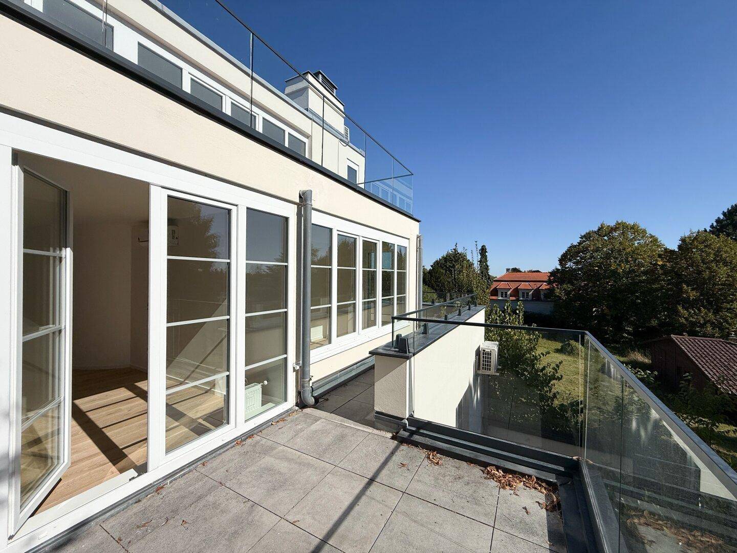 Helle Terrasse mit Fliesenboden, Glasgeländer und Zugang zu den Innenräumen des Gebäudes.