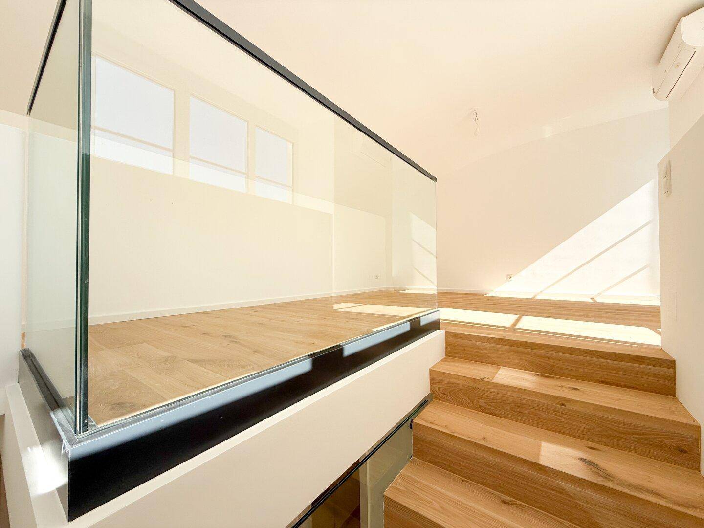 Moderne Holztreppe mit Glasgeländer, die zu einem hellen oberen Wohnbereich führt.