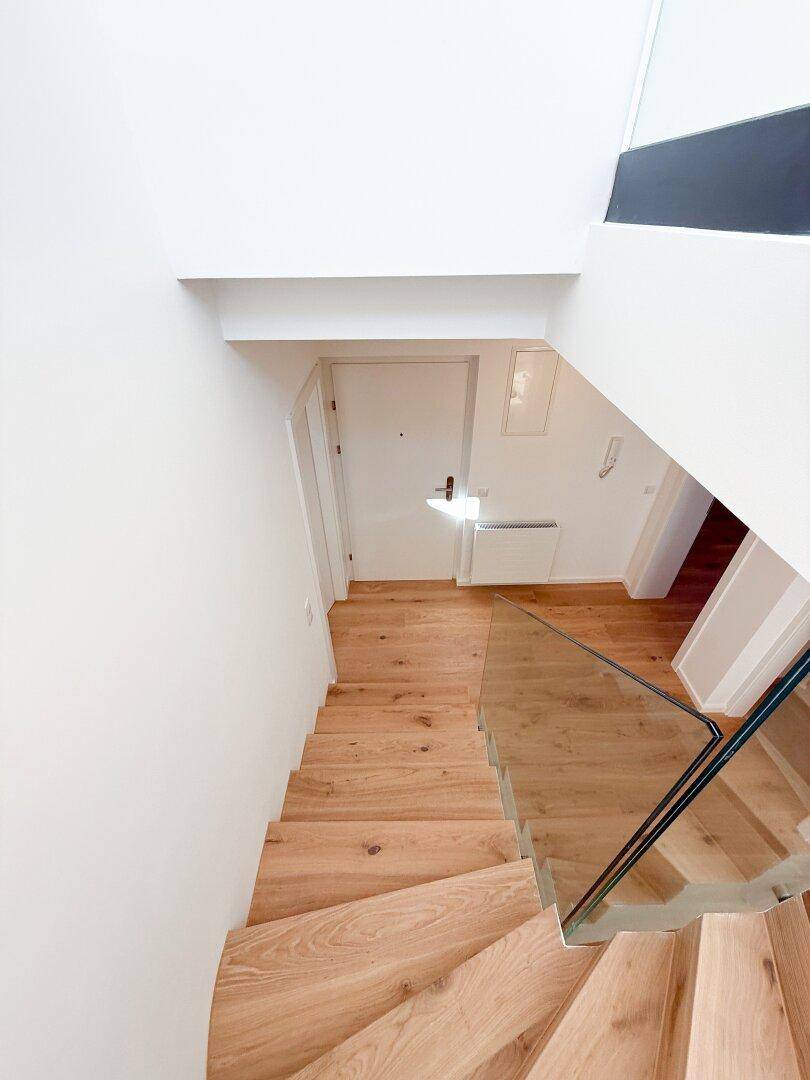 Blick von oben auf eine elegante Holztreppe mit Glasgeländer, die in den unteren Bereich führt.