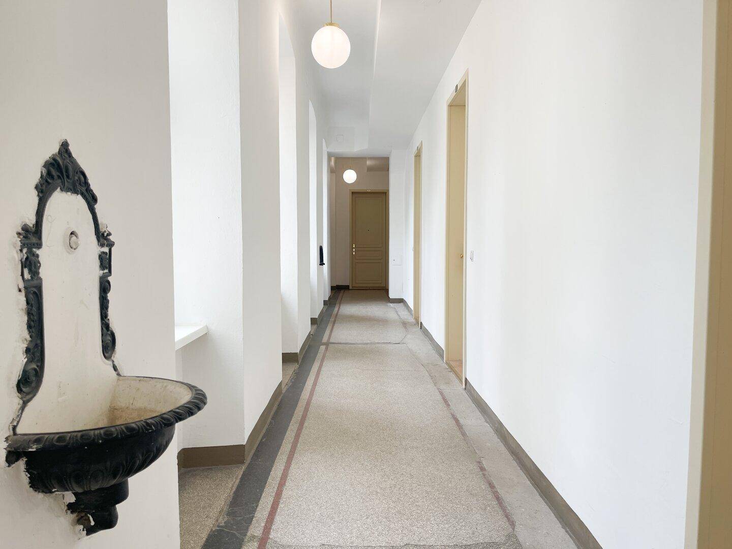 Langer Flur mit historischem Wandbrunnen und Terrazzoboden in einem Altbau mit hohen Decken.