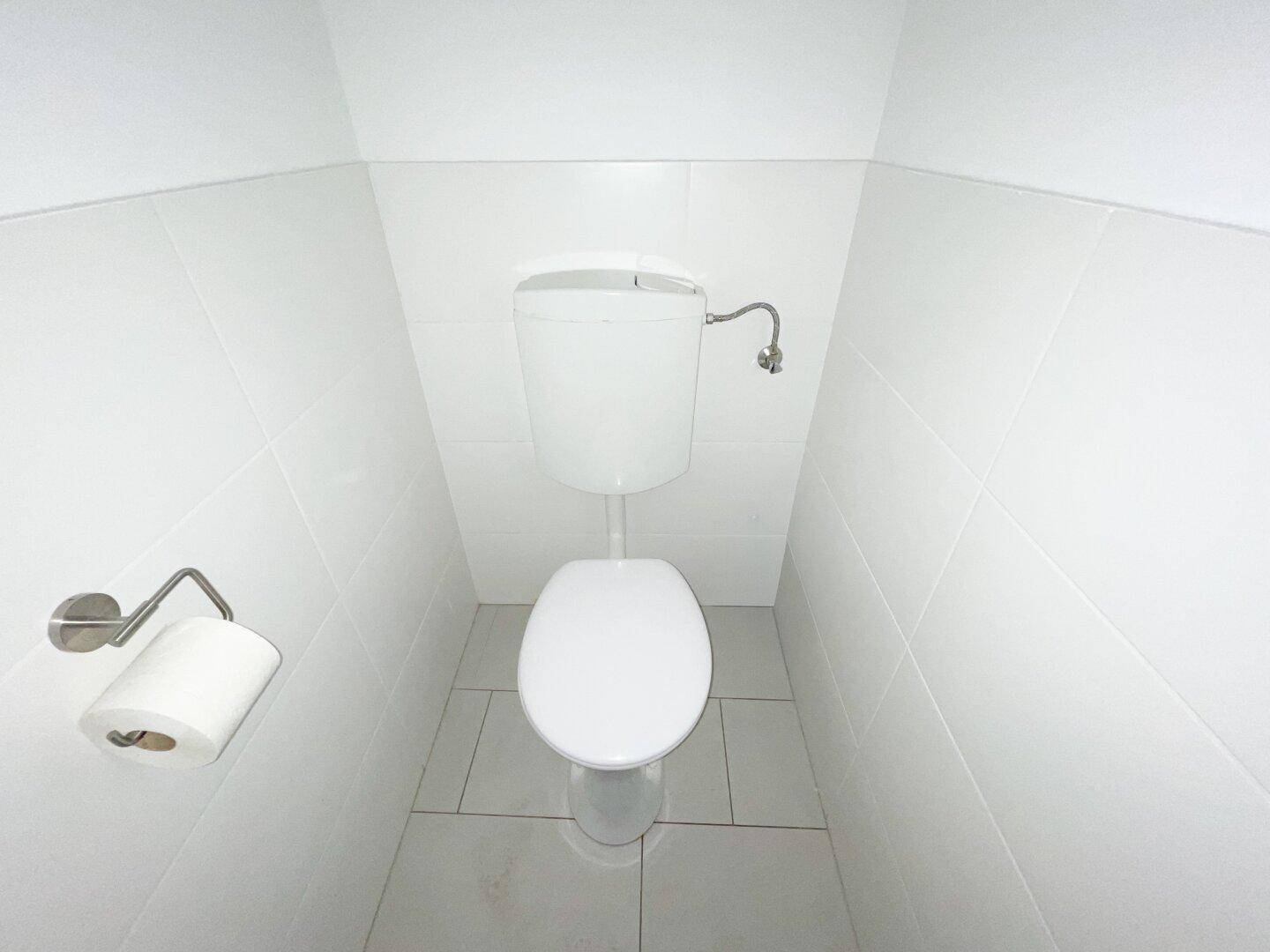 Eine kleine, helle Toilette mit weißen Fliesen an Wänden und Boden, ausgestattet mit einem einfachen WC.