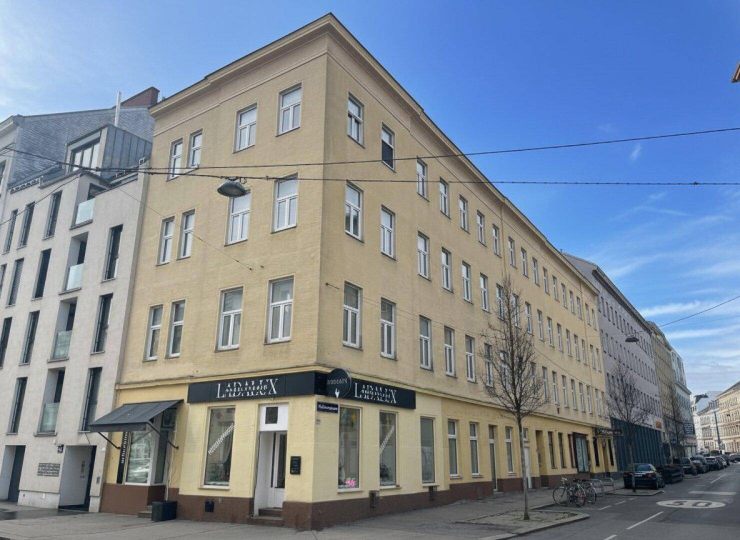 Ein Eckgebäude mit mehreren Stockwerken und Geschäftsflächen im Erdgeschoss, gelegen an einer städtischen Straße unter blauem Himmel.