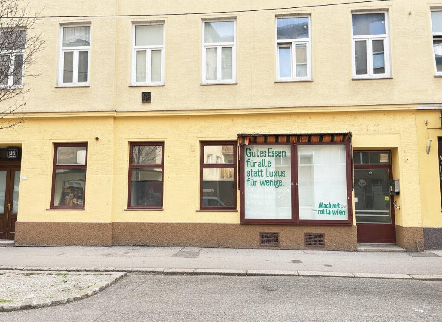 Eine breitere Ansicht der Ladenfront mit Schaufenstern und einer Werbebotschaft, eingebettet in die Fassade eines Mehrfamilienhauses.