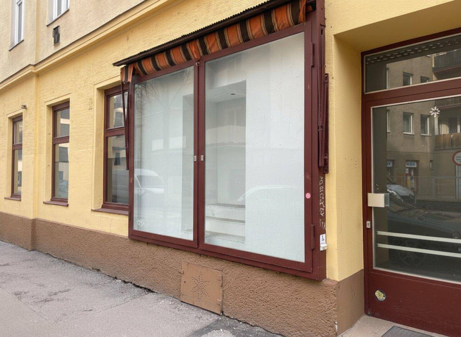 Eine Ladenfront mit großen Schaufenstern und einer Markise, Teil eines älteren Gebäudes an einer Straße.
