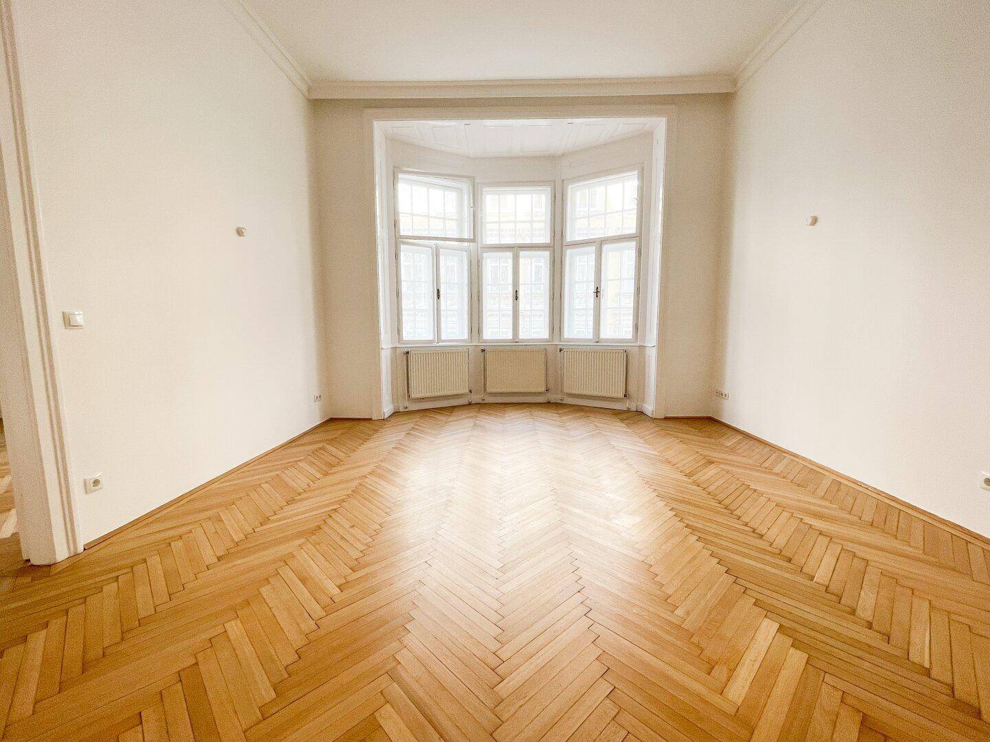 Geräumiges, leeres Zimmer mit klassischem Fischgrätparkett und einem breiten Erkerfenster.