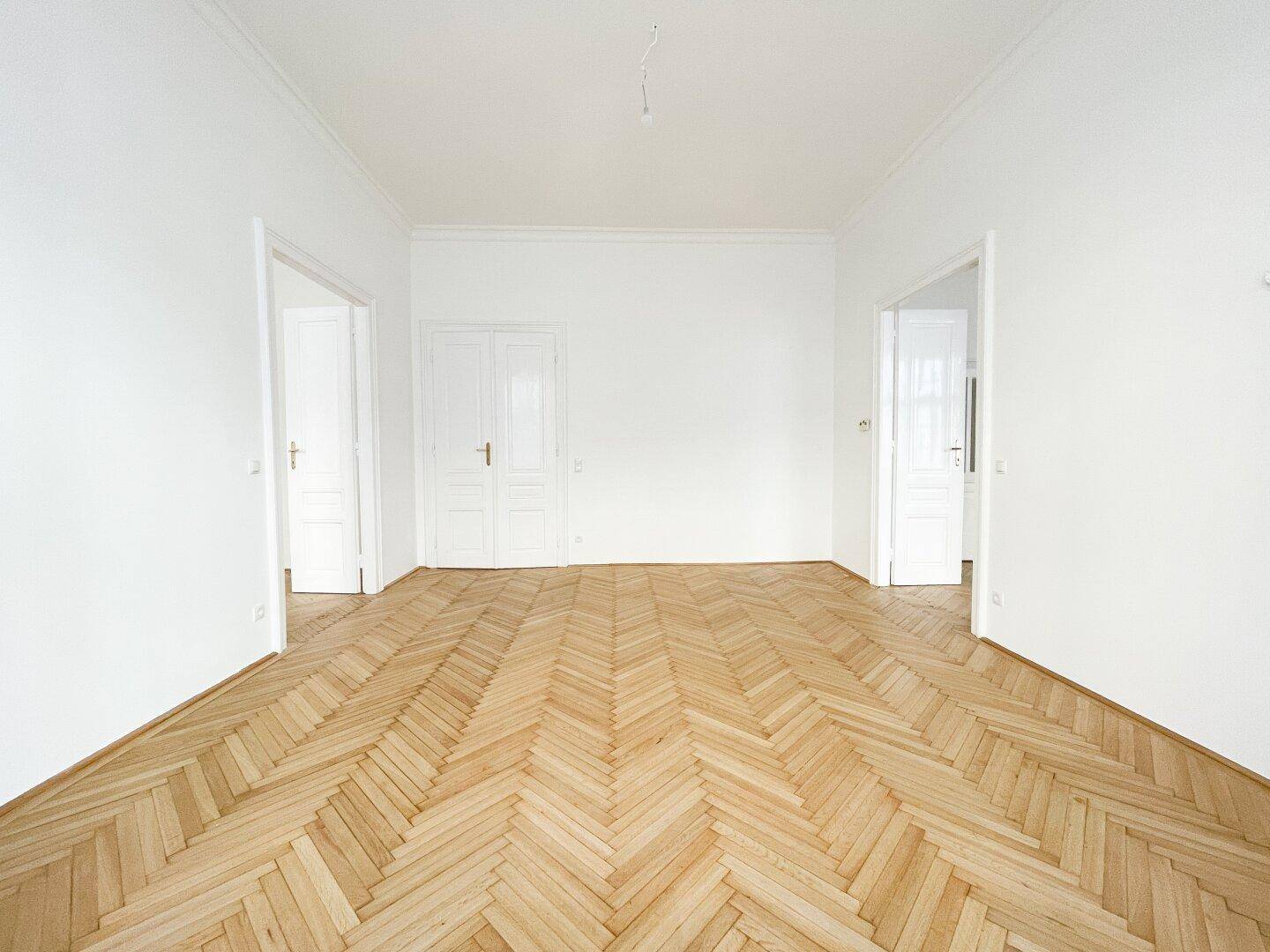 Leeres Zimmer mit Fischgrätparkett und zwei weißen Flügeltüren, die zu weiteren Räumen führen.