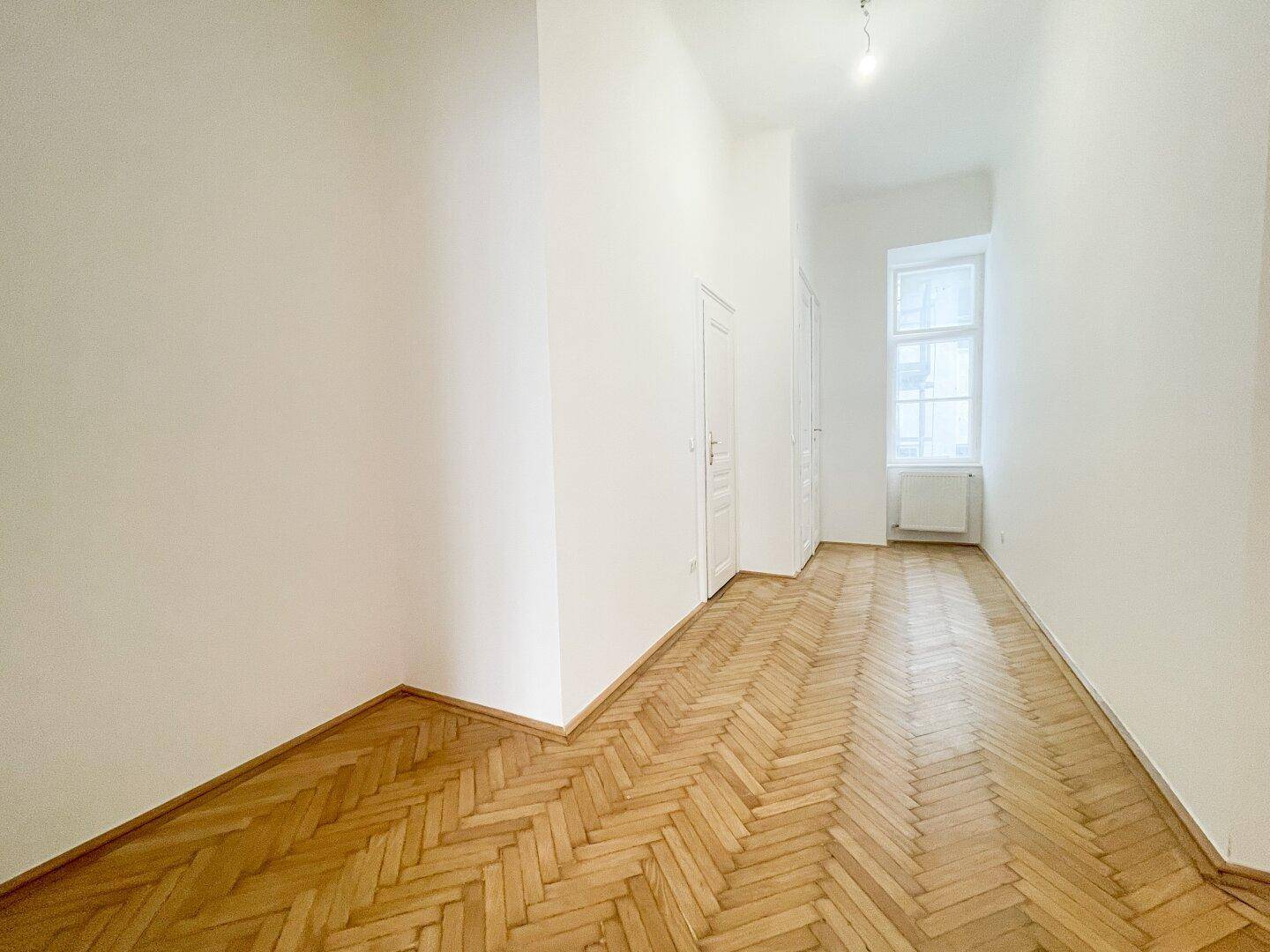 Langer Flur mit Fischgrätparkett und einem Fenster am Ende, der zu weiteren Zimmern führt.
