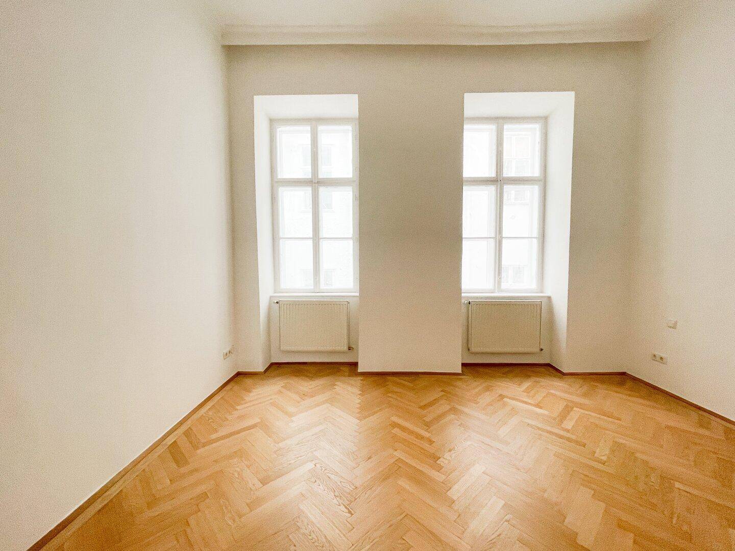 Leeres Zimmer mit Fischgrätparkett und zwei hohen Fenstern, die für natürliche Belichtung sorgen.
