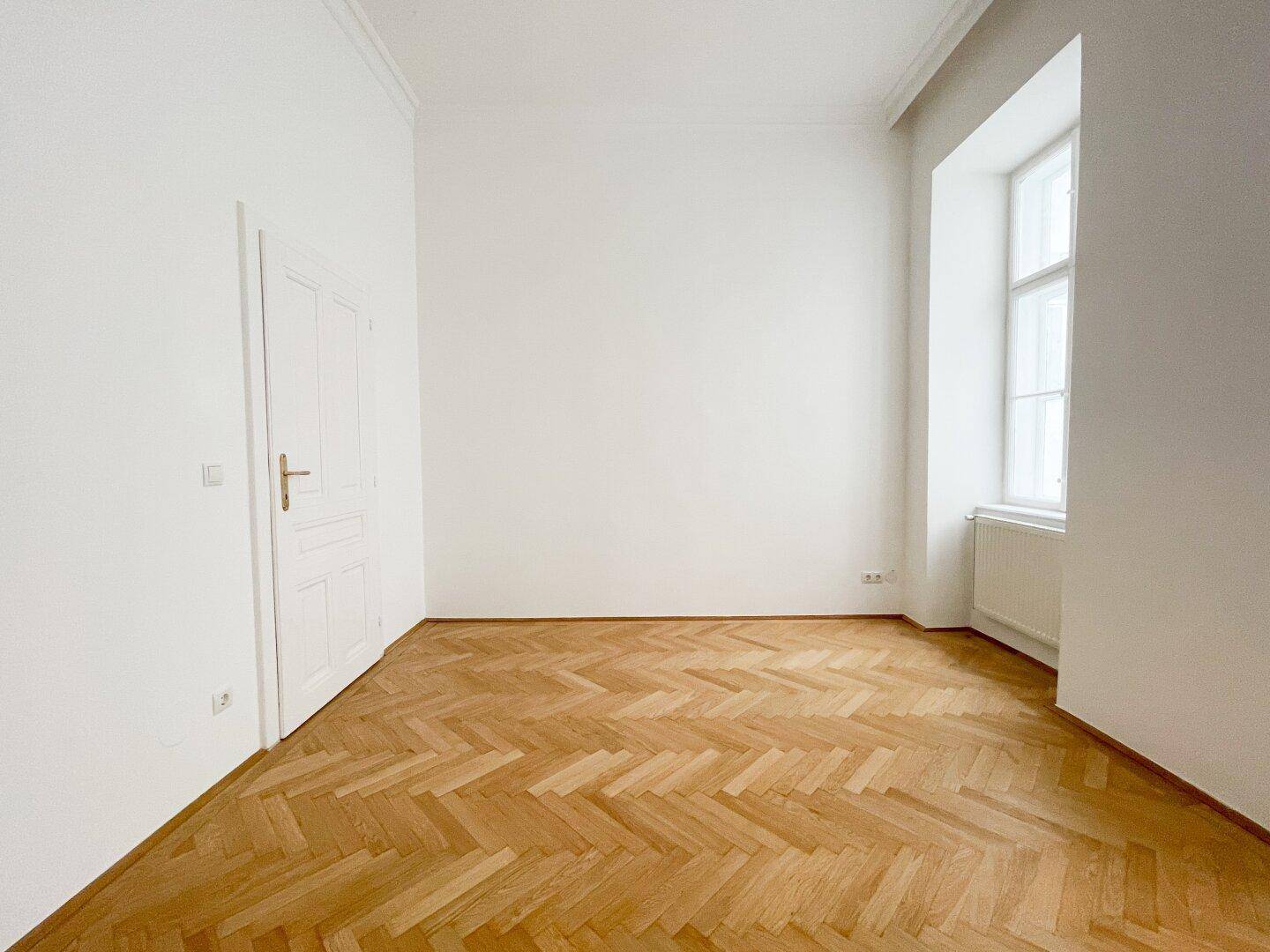 Leeres Zimmer mit Fischgrätparkett und einem Fenster, das für eine angenehme Helligkeit sorgt.