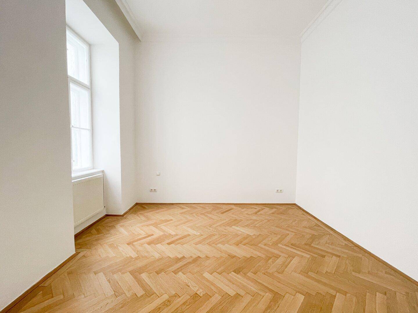 Heller, leerer Raum mit Fischgrätparkett und einem Fenster, das den Raum mit Tageslicht füllt.
